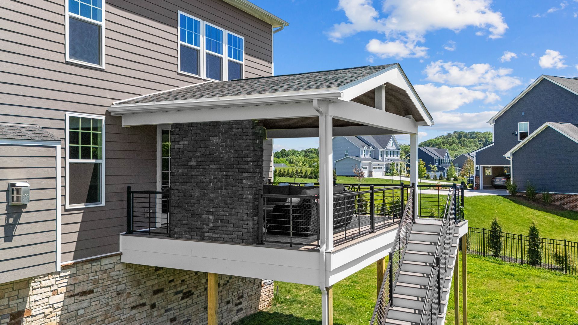 Covered backyard deck with gray siding, white trim, and stairs beside a house on a sunny day.