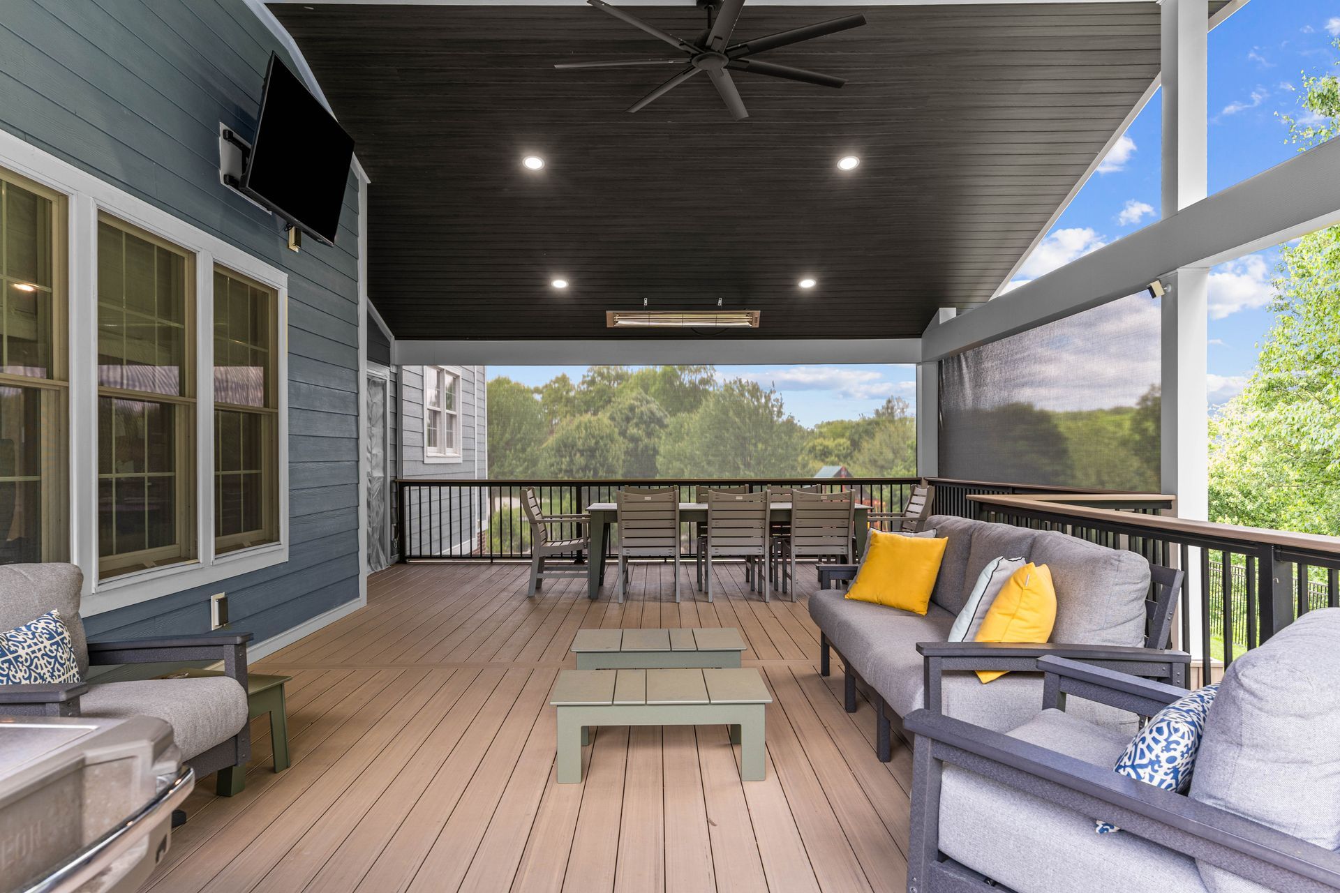 Covered outdoor porch with gray seating, yellow pillows, and dining table overlooking trees