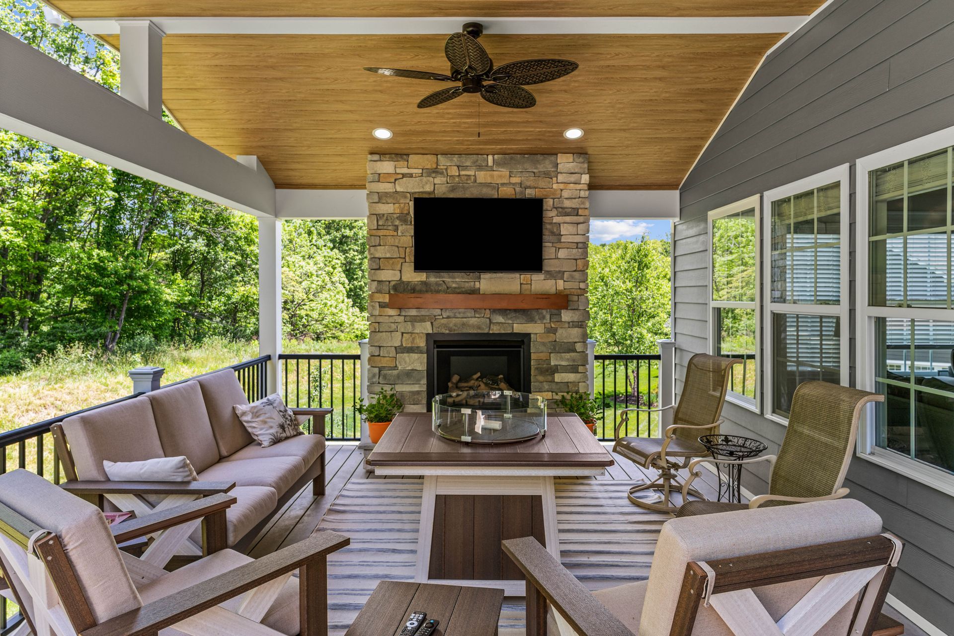 Covered patio with cushioned chairs, stone fireplace, wall TV, and wood ceiling overlooking greenery