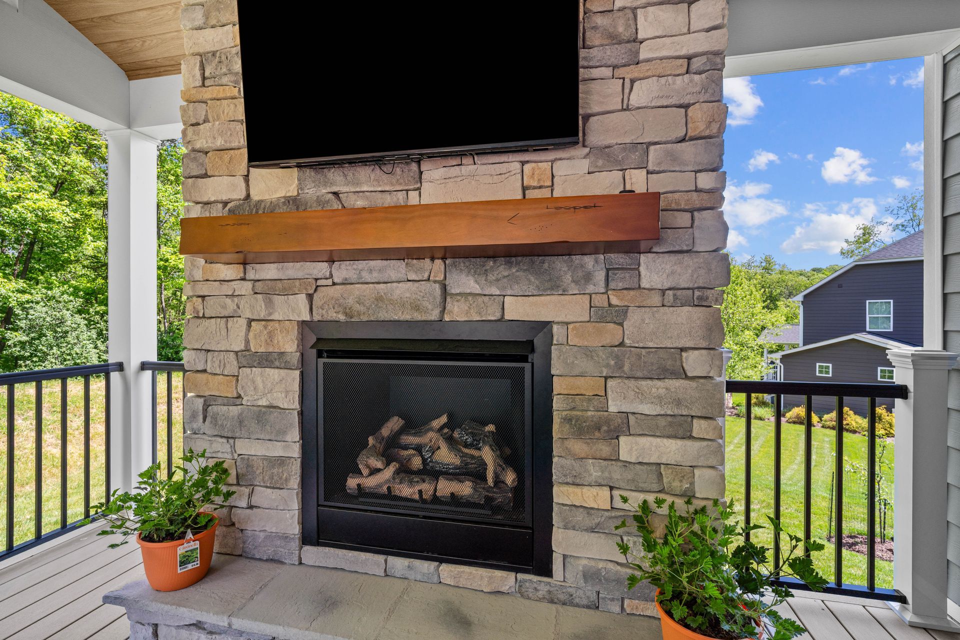 Covered porch with stone fireplace, mounted TV, and potted plants overlooking a green yard.