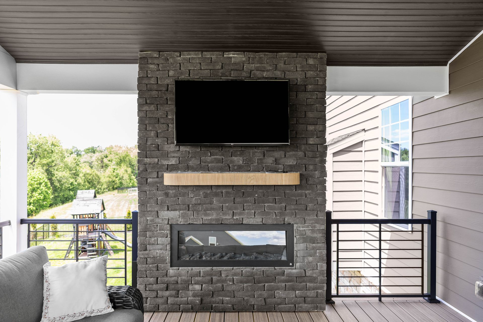 Covered balcony with a stone fireplace, TV, and railing, overlooking trees and neighboring buildings.