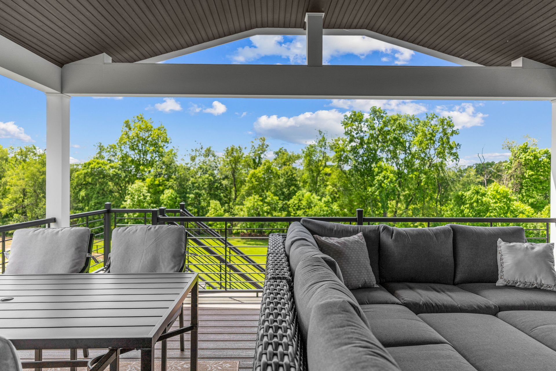 Covered patio with gray outdoor seating and table overlooking green trees and blue sky