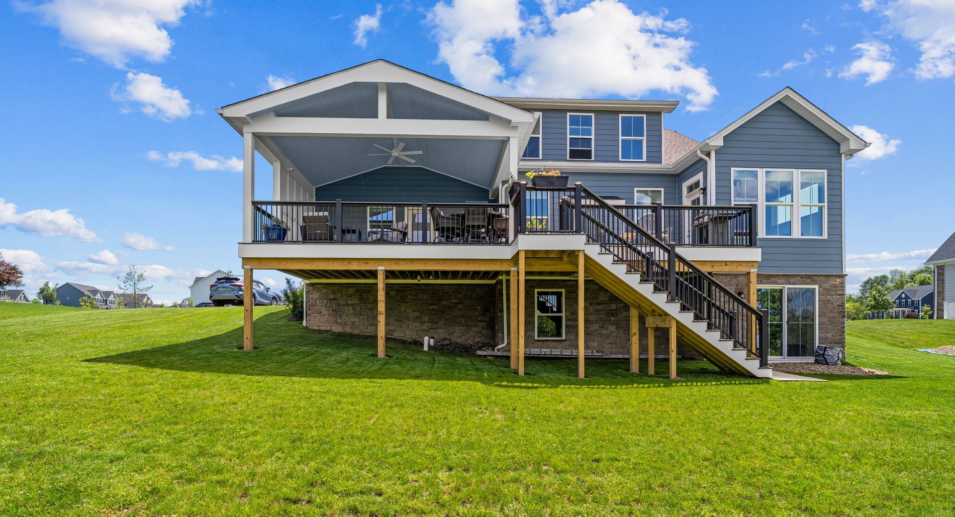 Raised gray house with deck and stairs on a grassy hillside under a blue sky