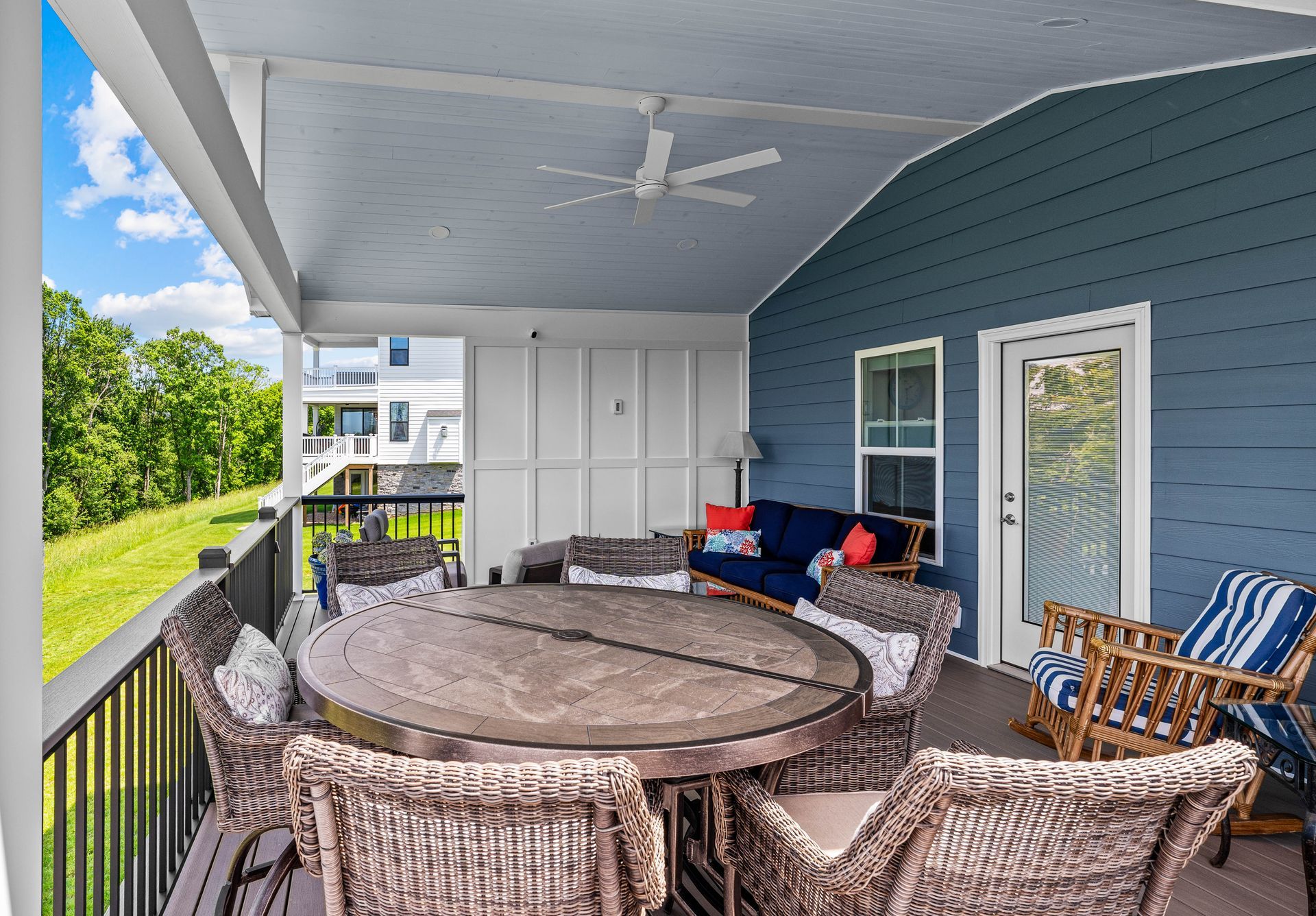Covered porch with wicker seating around a round table, blue siding, and a ceiling fan overlooking greenery.