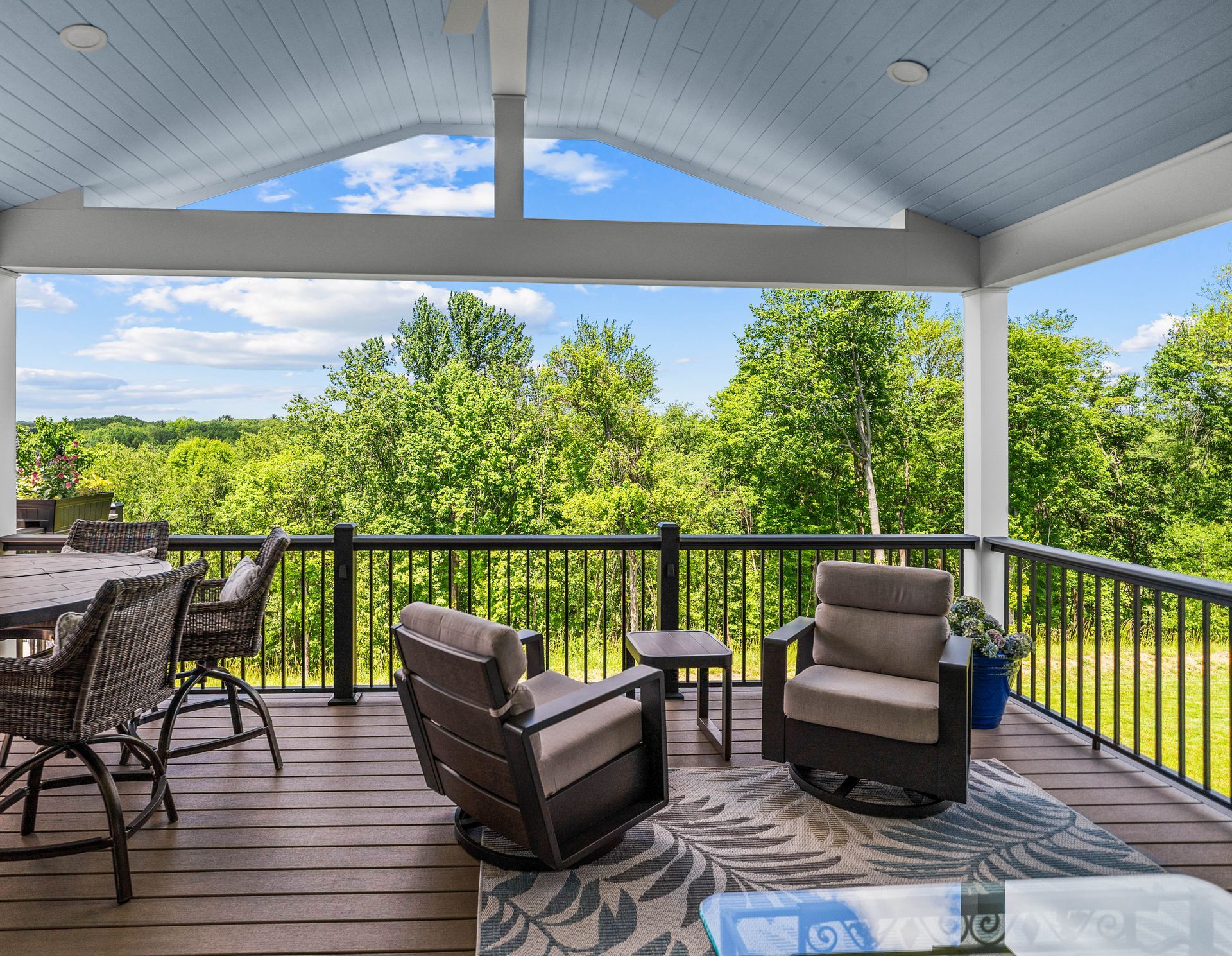 Covered wooden porch with cushioned chairs, glass table, and a green wooded view beyond the railing