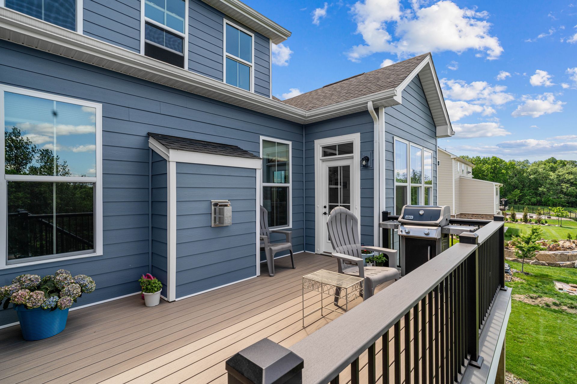 Blue house exterior with a small deck, black railing, potted plants, and lawn under a sunny sky