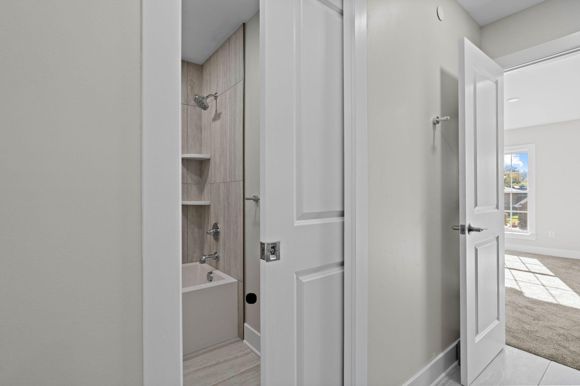 Hallway view of a bright bathroom with white doors and gray tile, leading to a sunlit bedroom.