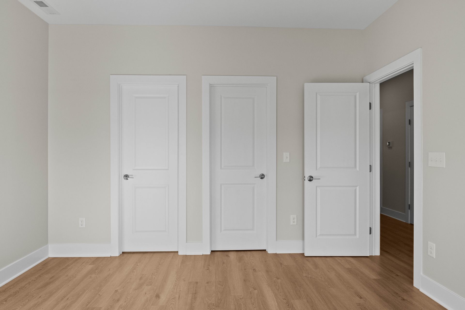 Empty beige room with hardwood floor and four white doors, one open to a hallway