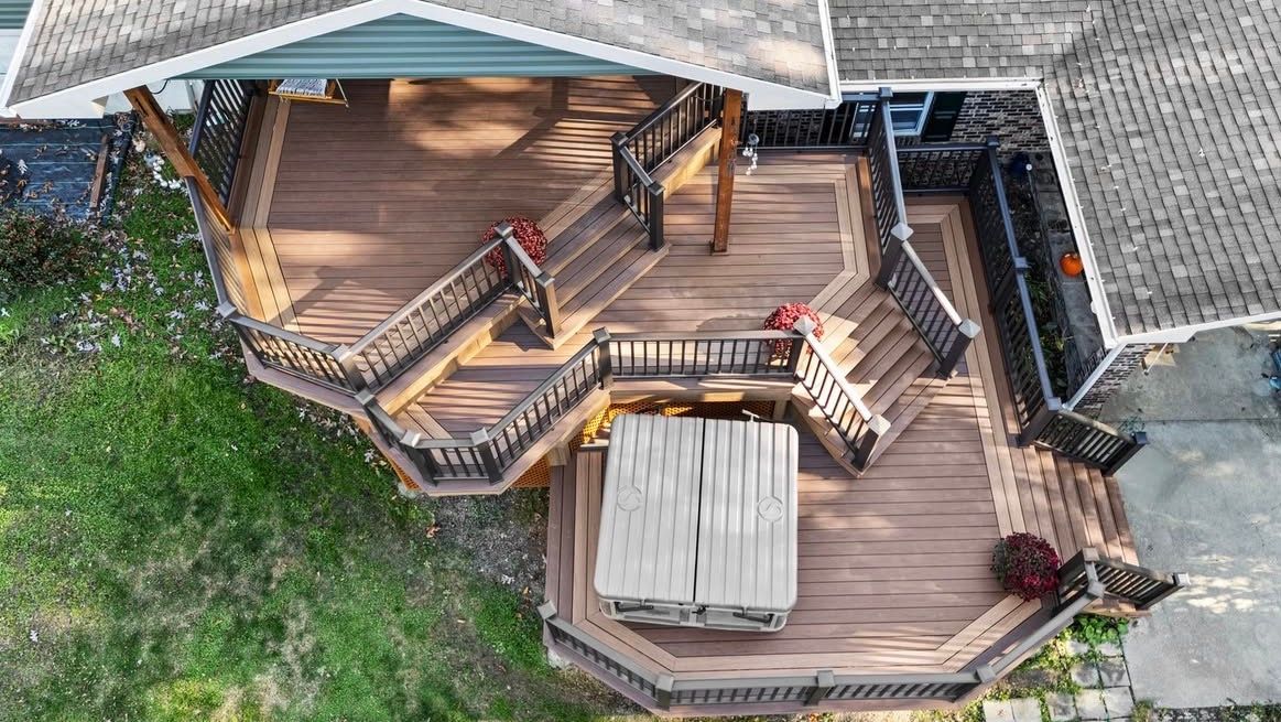 Aerial view of a multi-level wooden deck with stairs and a central hot tub. Green grass surrounds.