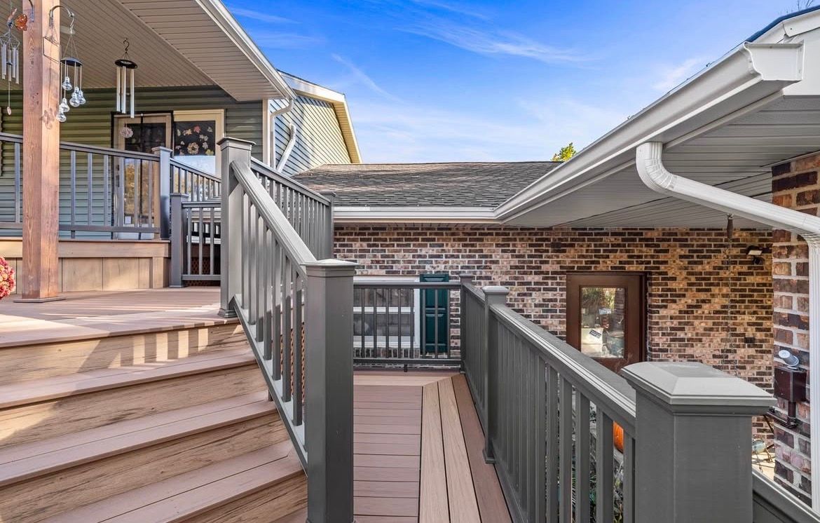 Exterior view of a house with a staircase and a deck, gray railing, brown wood, brick wall, and blue sky.