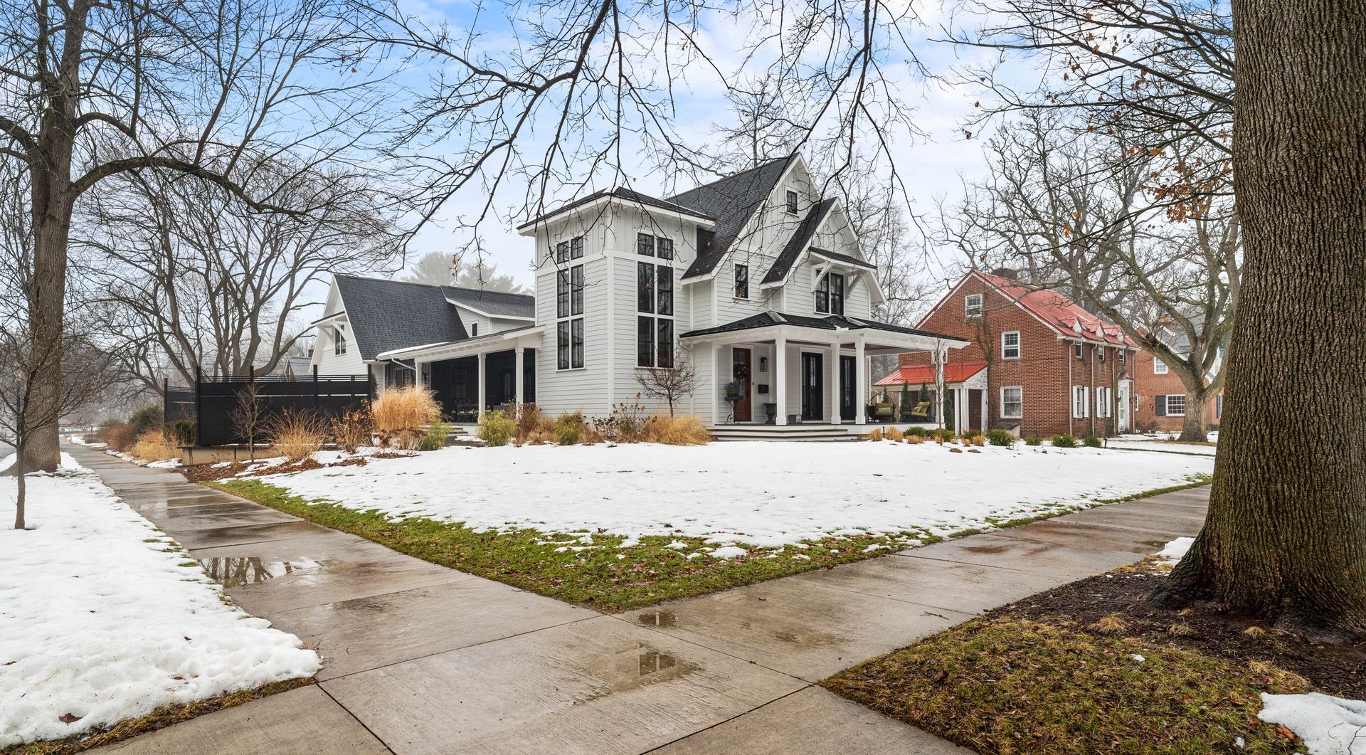 Snowy suburban house with a wraparound porch and bare trees beside a sidewalk