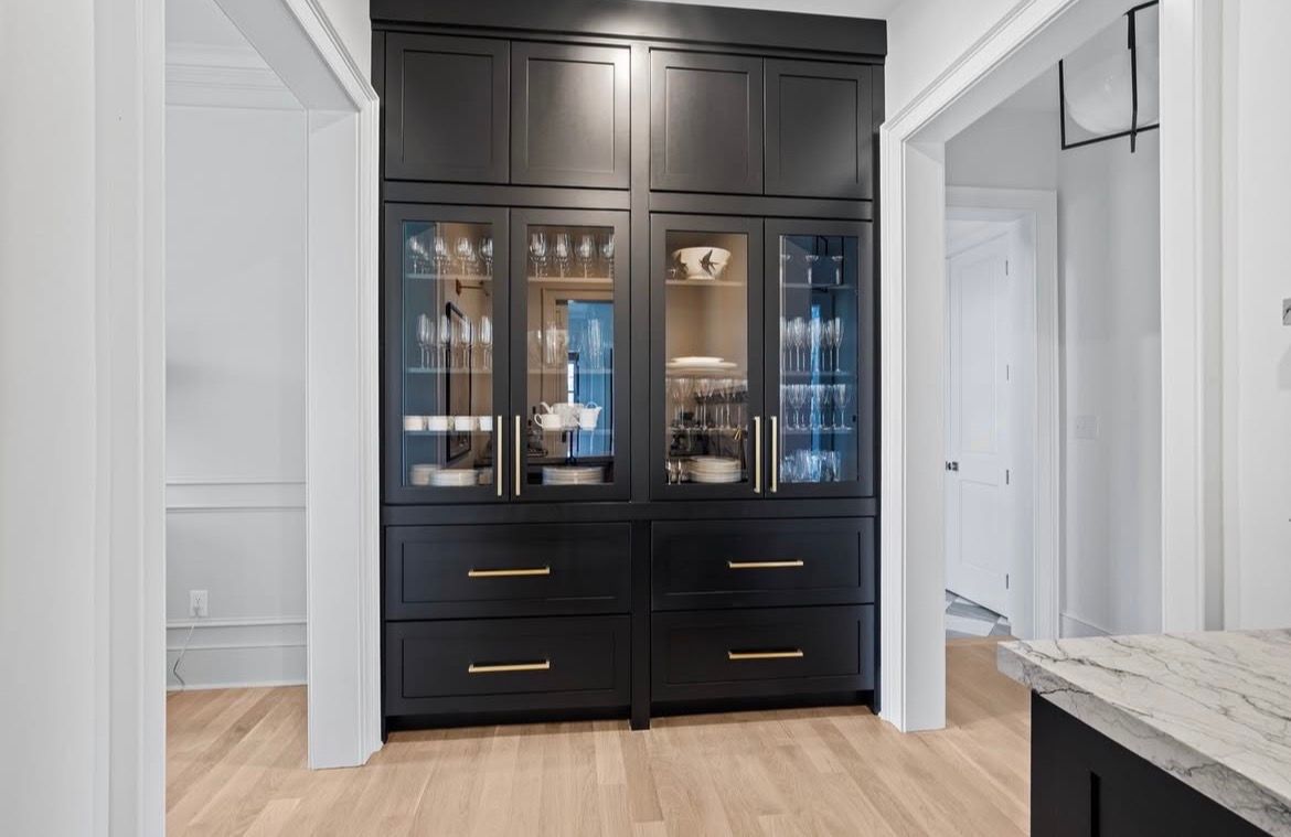 Black kitchen cabinet with glass doors, gold hardware, and drawers; installed in a hallway with wooden floors.