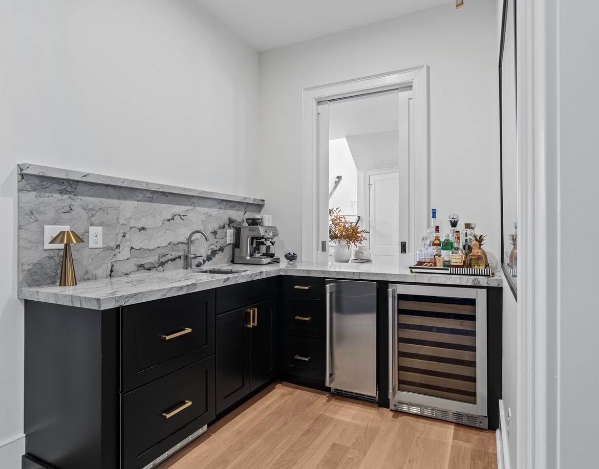 L-shaped home bar with black cabinets, gray countertops, stainless steel appliances, and a window.