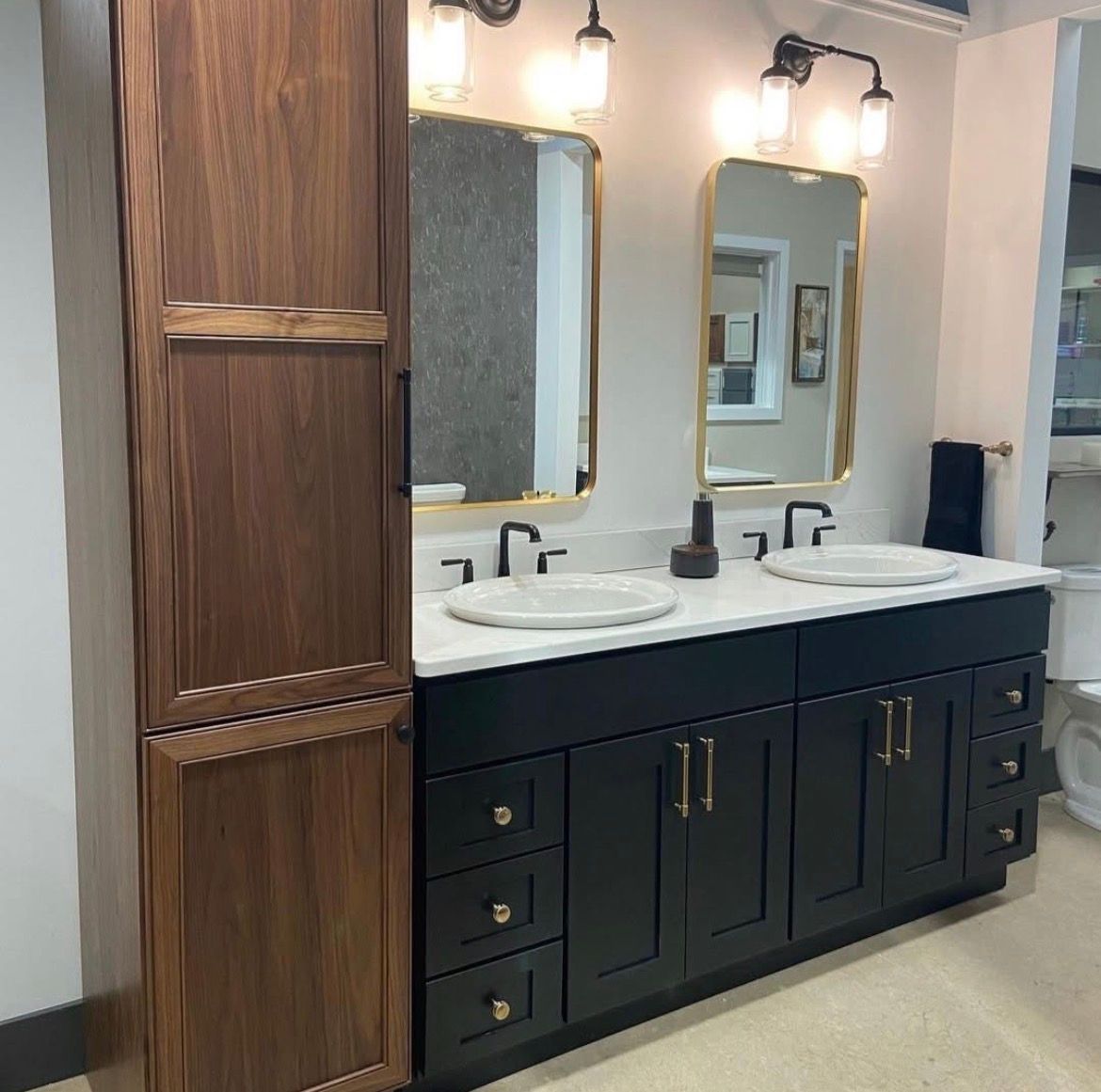 Bathroom with black vanity, gold mirrors, white countertop, walnut storage cabinet, and sconces.