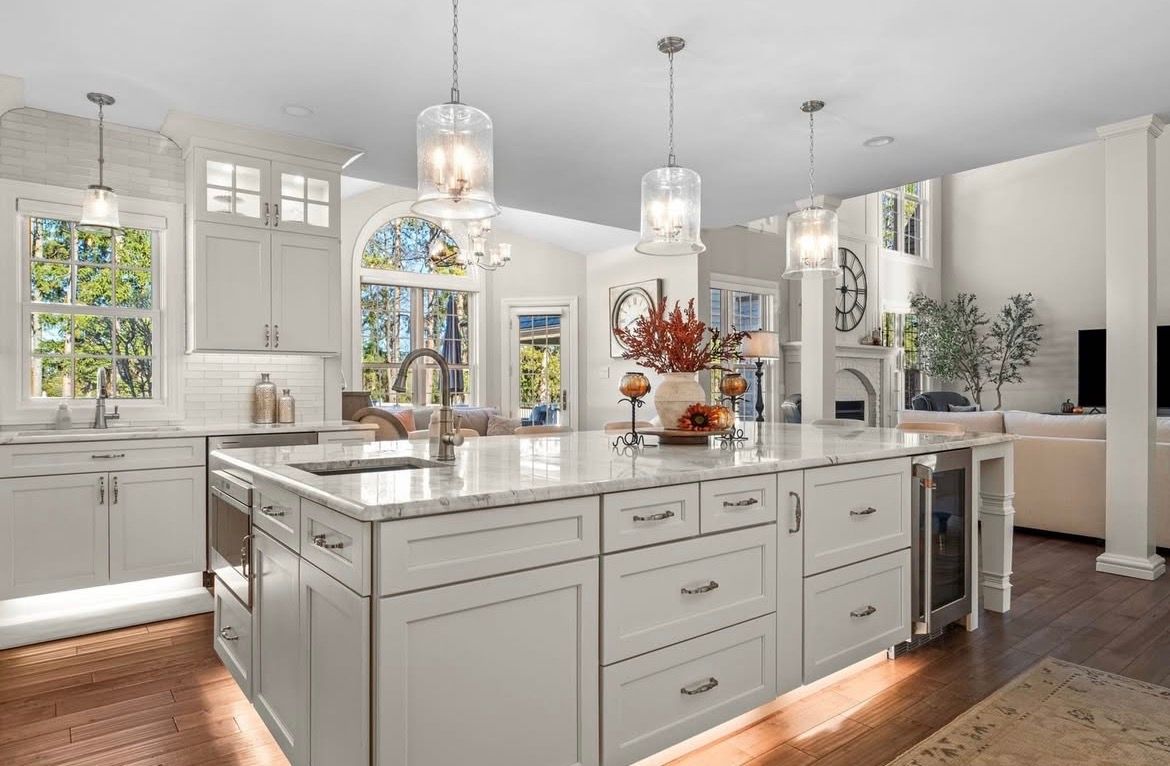 White kitchen with large island, marble countertops, pendant lights, and hardwood floors.