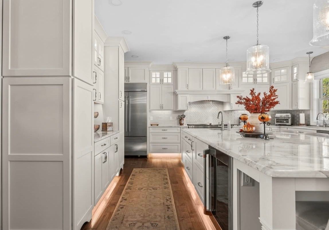 Bright white kitchen with large island, stainless steel refrigerator, and wooden floors.