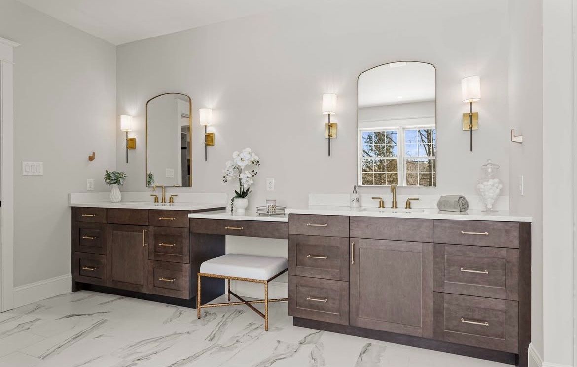 Bathroom with two vanities, white countertops, dark wood cabinets, arched mirrors, gold sconces, white stool.