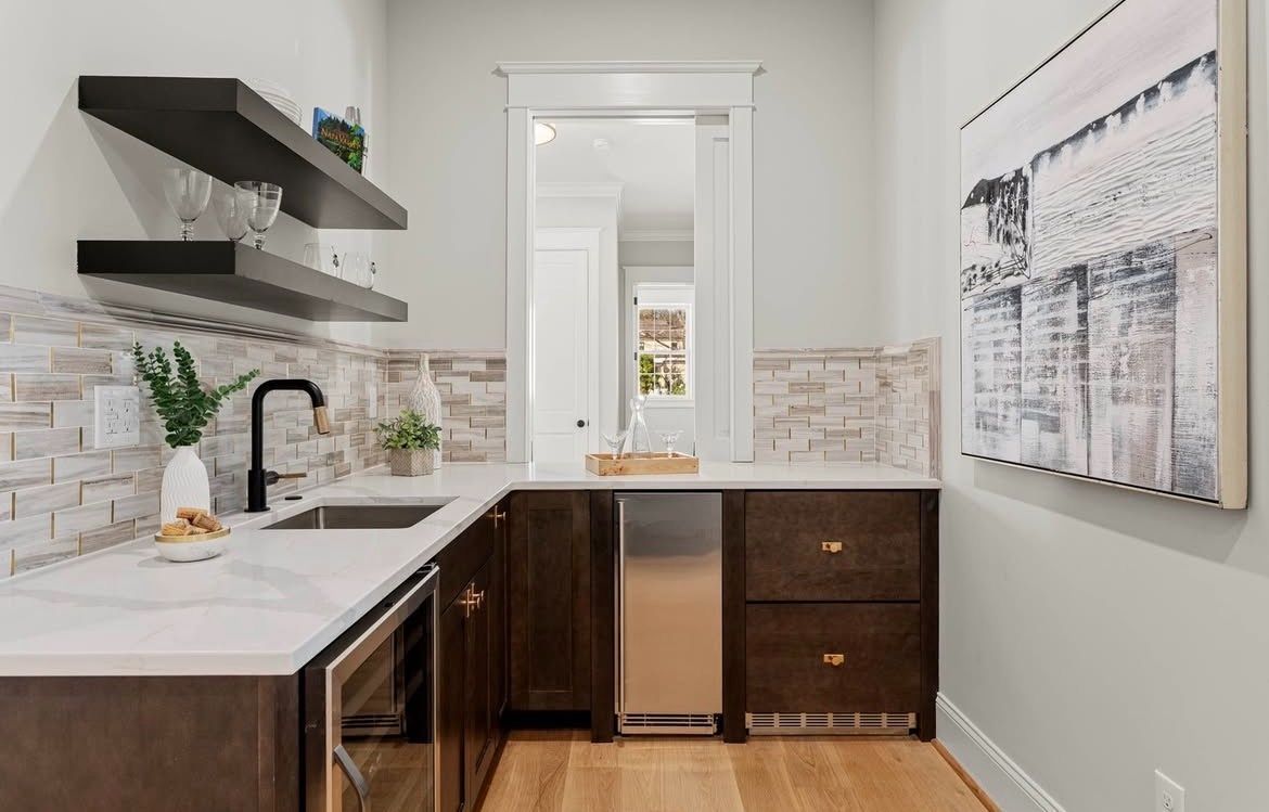 A home bar area with white countertops, dark cabinets, and a brick-look backsplash, with floating shelves.