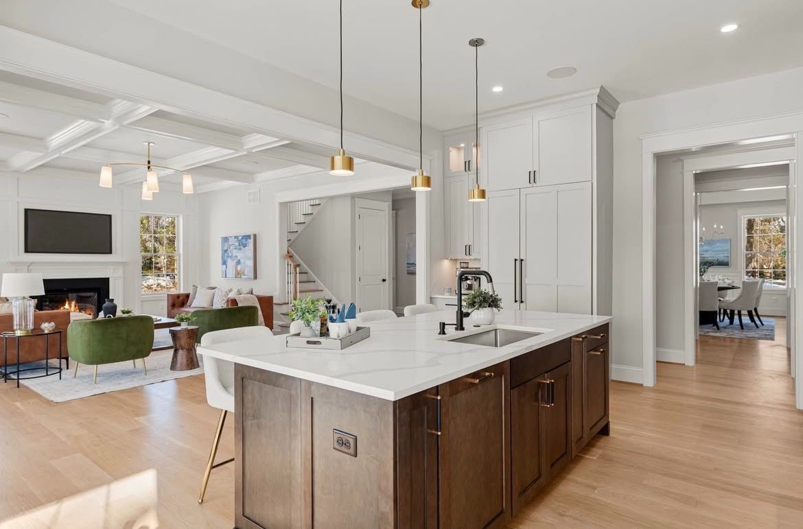 Modern kitchen with island, adjacent living room, and a staircase in the background. White and brown color scheme.