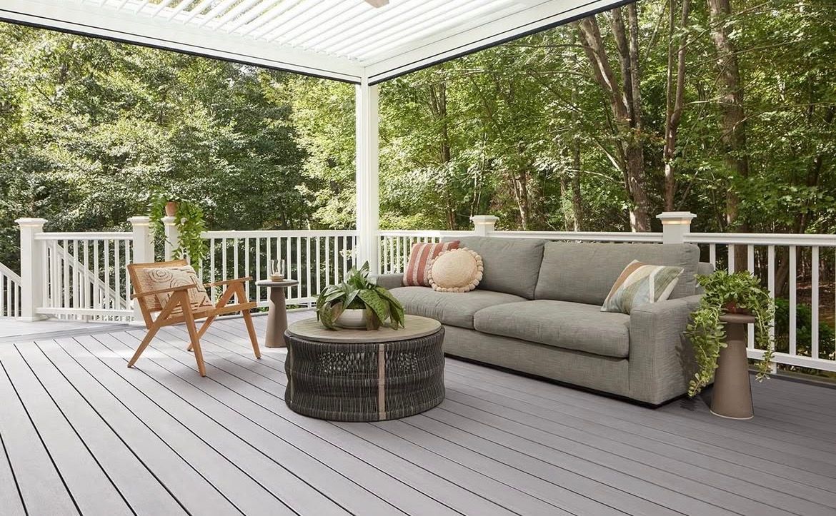 Covered porch with gray decking, white railing, and a gray sofa with colorful pillows; trees in the background.