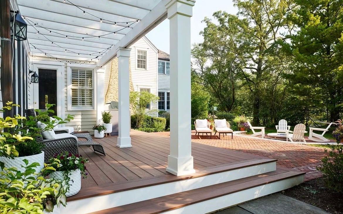 White-columned outdoor patio with seating, steps, and a pergola; a brick patio and a white house with greenery.