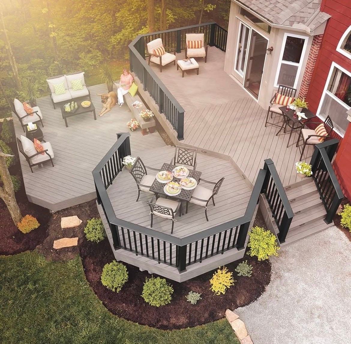 Overhead view of a multi-level deck with seating, next to a red house. Gray decking, dark railings, and a green yard.