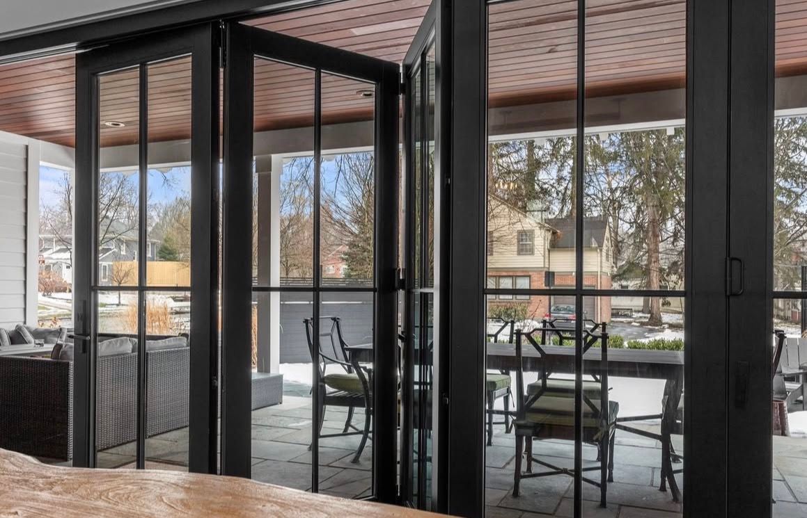 Black-framed glass doors open to outdoor dining area with wrought iron chairs and table. View of trees and house beyond.