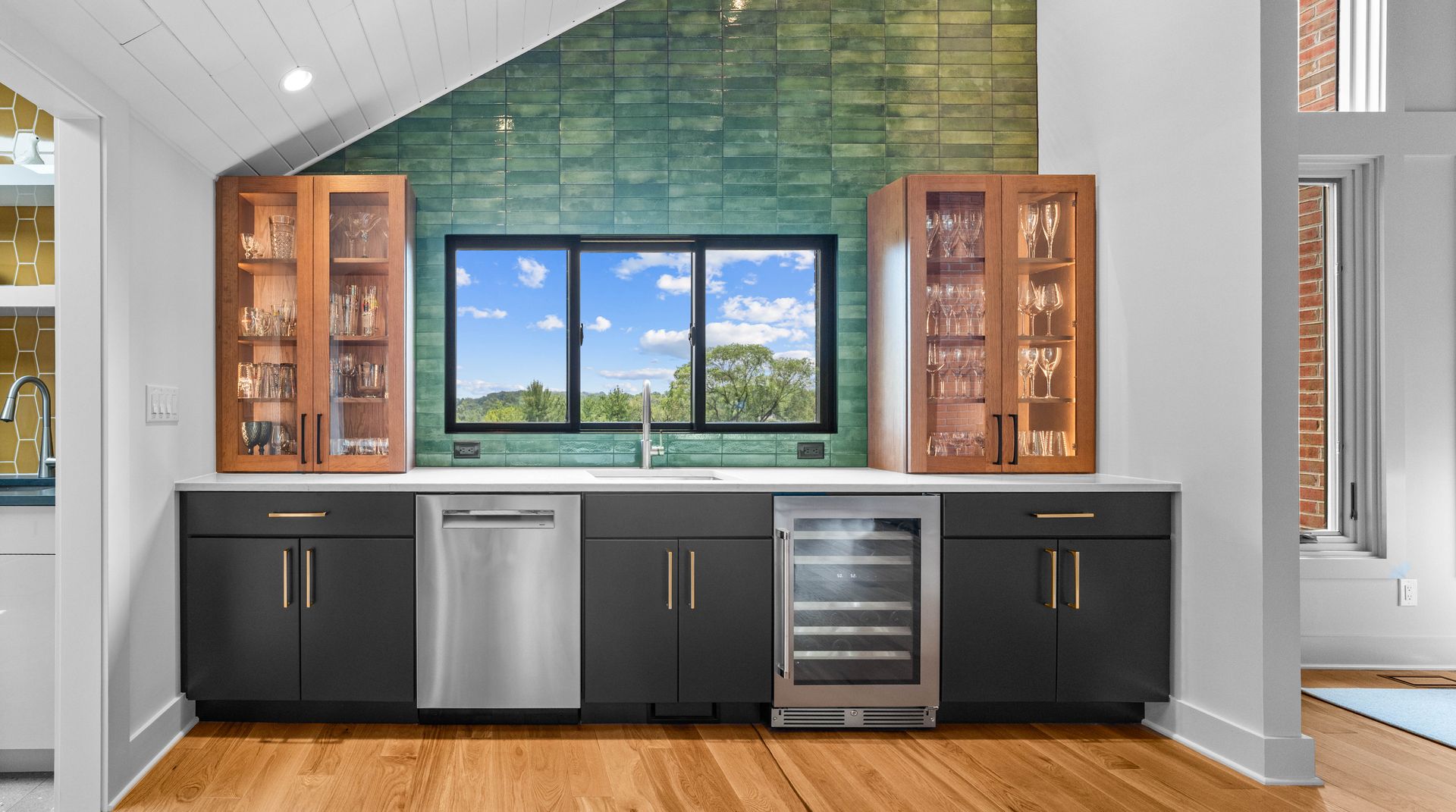 Modern outdoor kitchen with black cabinets, stainless appliances, wood accents, and a window view to blue sky.
