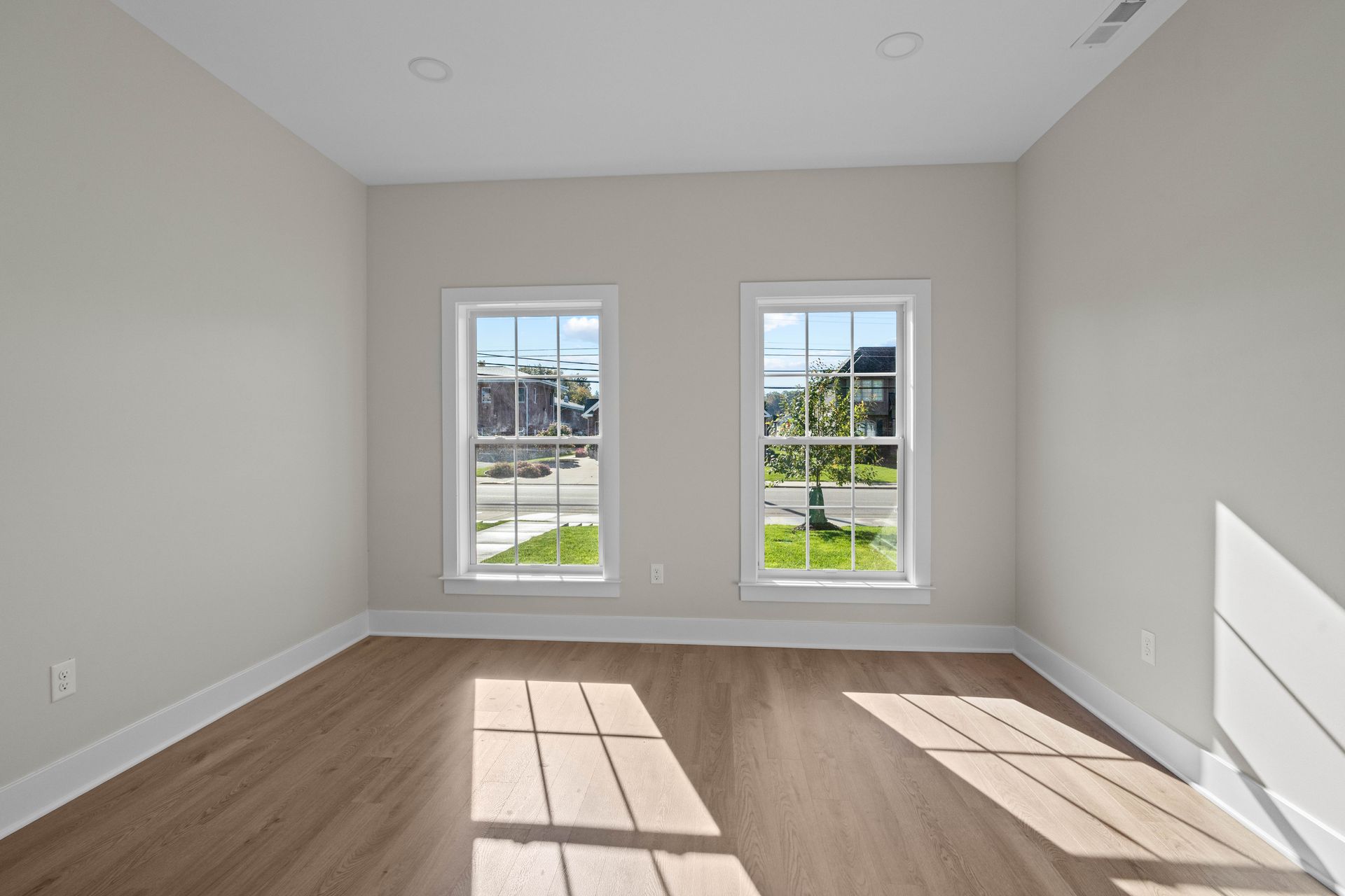 Empty sunlit room with two windows, light wood floor, and white walls
