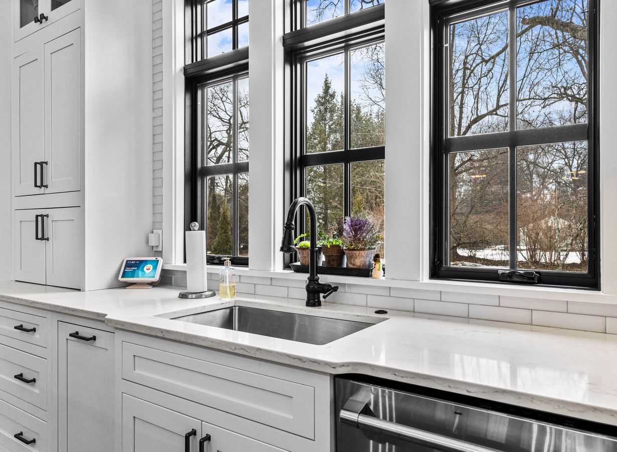 Bright white kitchen with large windows, a marble countertop, and a stainless sink overlooking trees outside