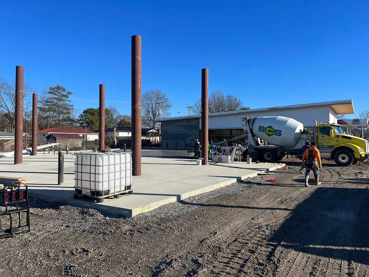 A yellow cement truck is driving down a dirt road in front of a building under construction.