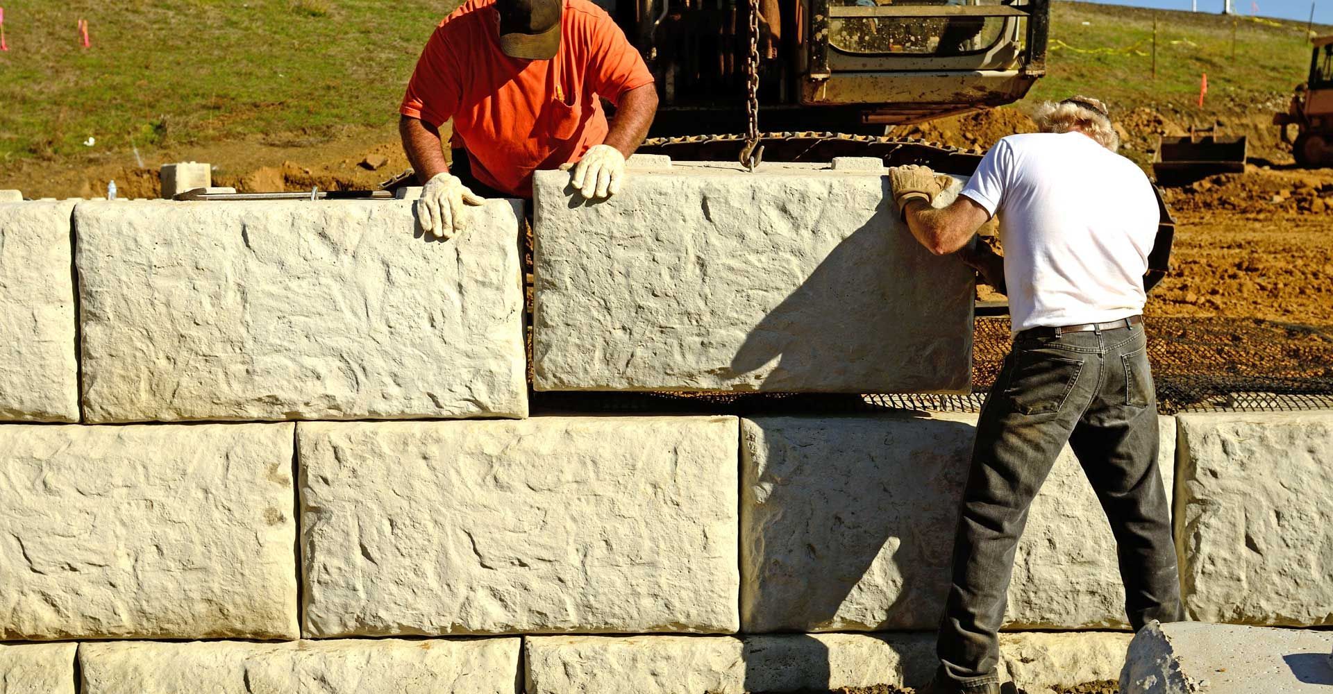 Two men are working on a stone wall with a bulldozer in the background.