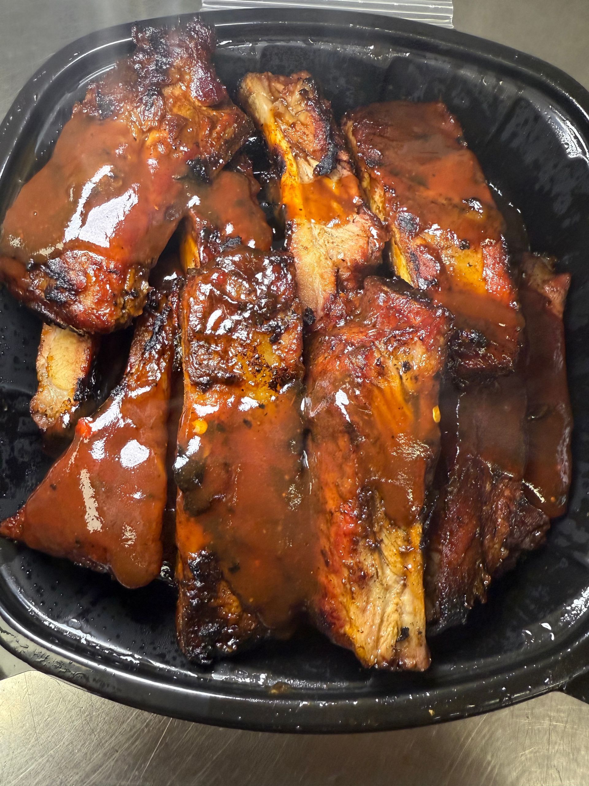 Several cooked pork ribs coated in a thick, glossy, reddish-brown barbecue sauce served in a black plastic container.