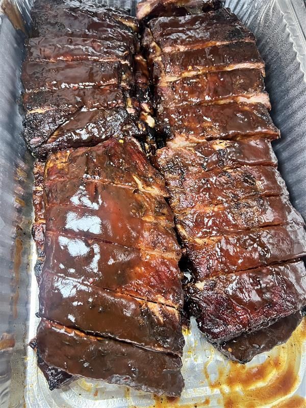 Two racks of glazed, dark-brown barbecued ribs cut into individual portions inside a rectangular aluminum foil tray.