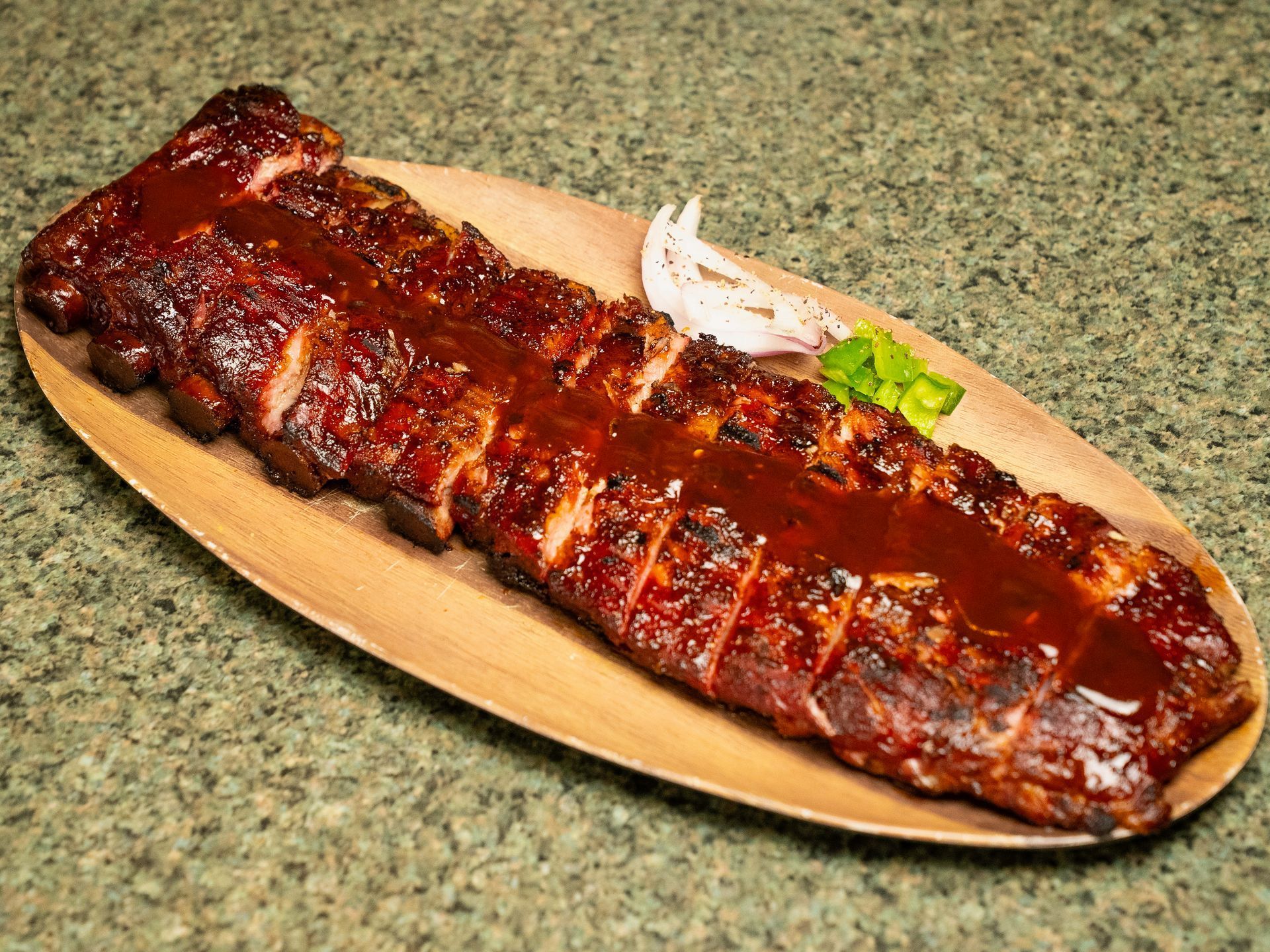 A rack of sliced, barbecue-glazed pork ribs served on an oval wooden platter with a side of white onions and herbs.