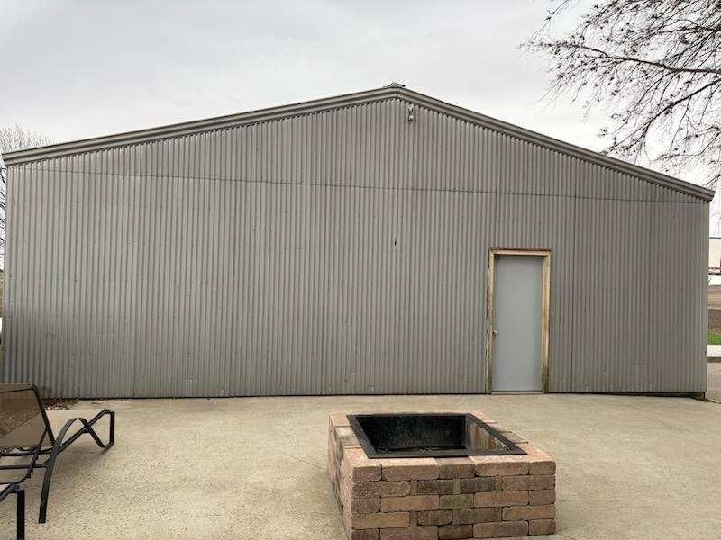 Gray corrugated metal building with a door, fire pit, and gravel patio. Overcast sky.