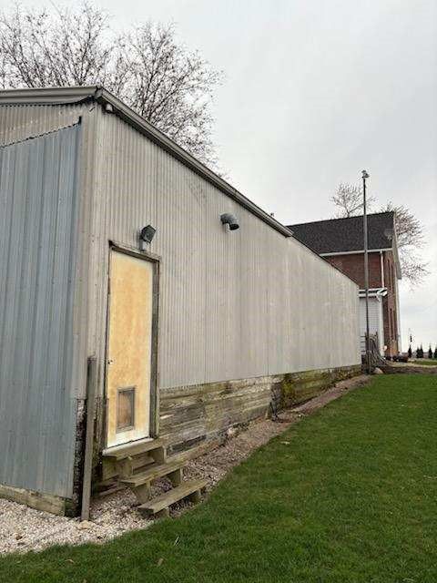 Exterior of a long, gray metal-sided building with a door, steps, and a grassy area in front.