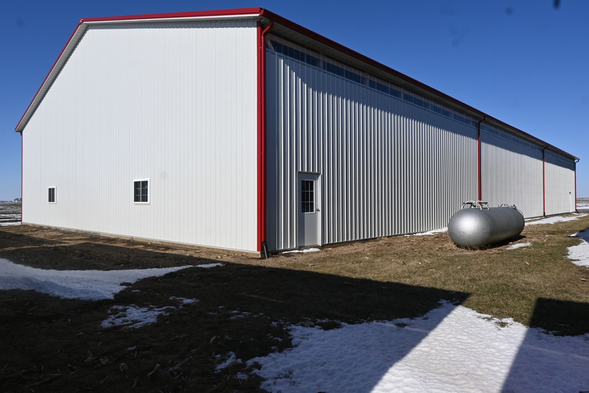 White metal barn with red trim, door, and a propane tank. Snowy ground.