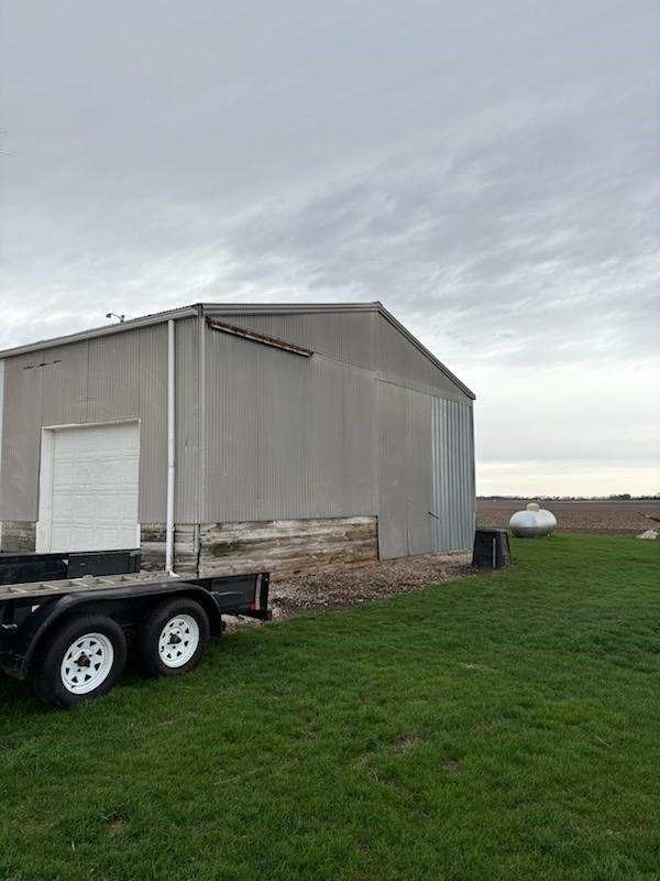 A metal shed with a white door, trailer, and propane tank on green grass under a cloudy sky.