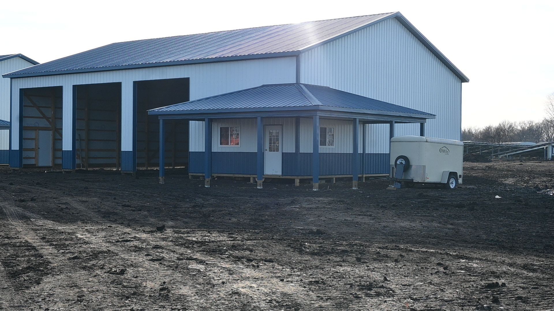 Large white warehouse with a small blue and white shed in front; muddy ground.