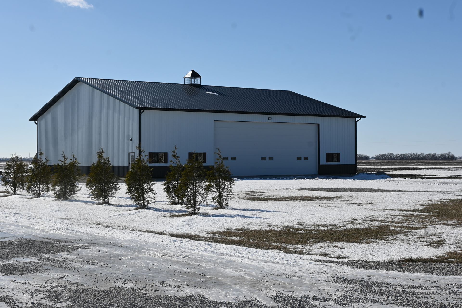 White metal barn with a black roof, in a snowy field on a sunny day.