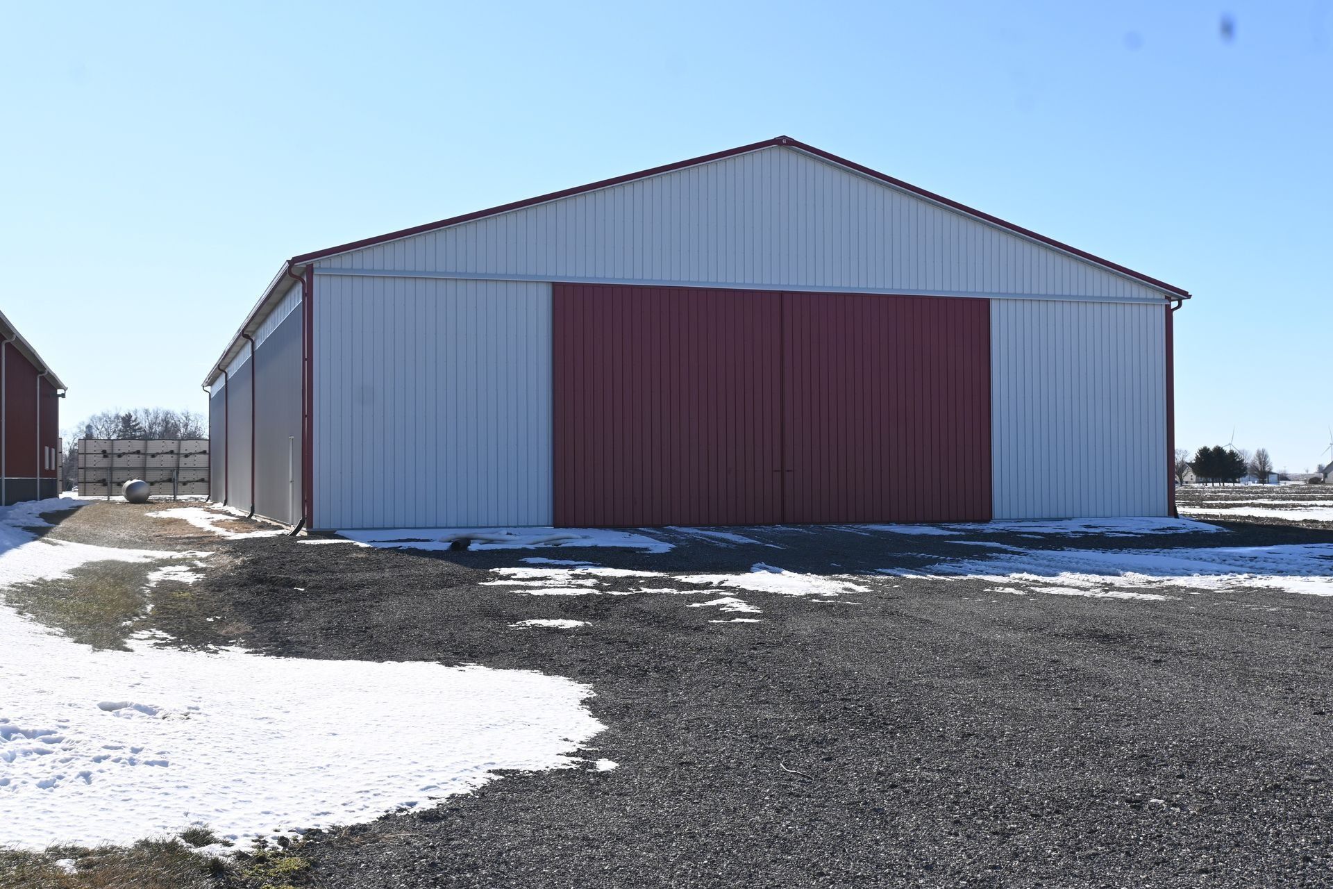 White and red metal barn with a gravel driveway and patches of snow on a sunny day.