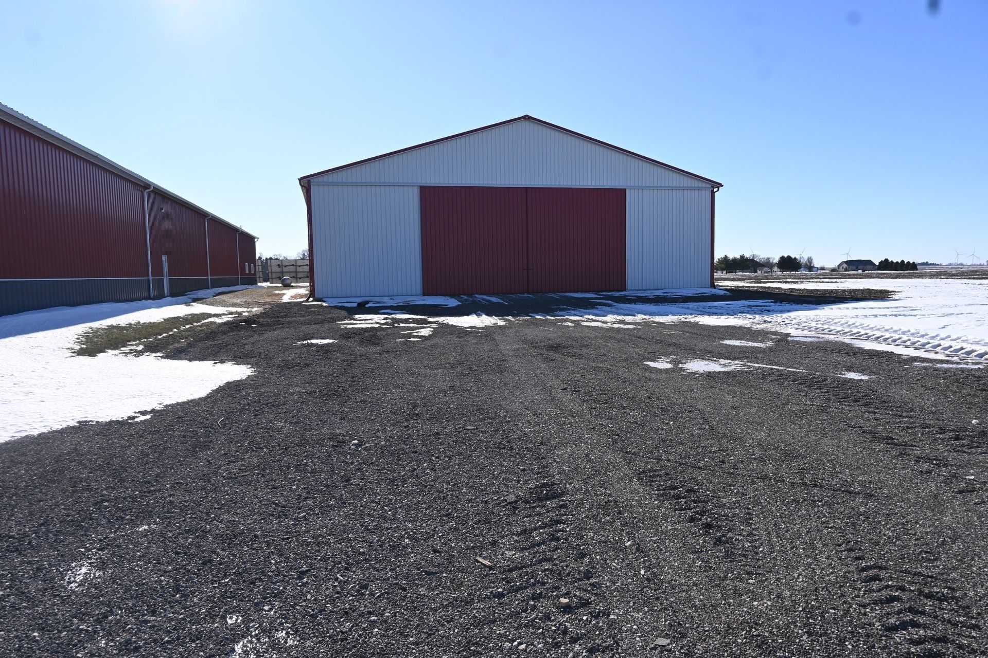 A white metal barn with red doors and roof on a gravel surface, surrounded by snow patches.