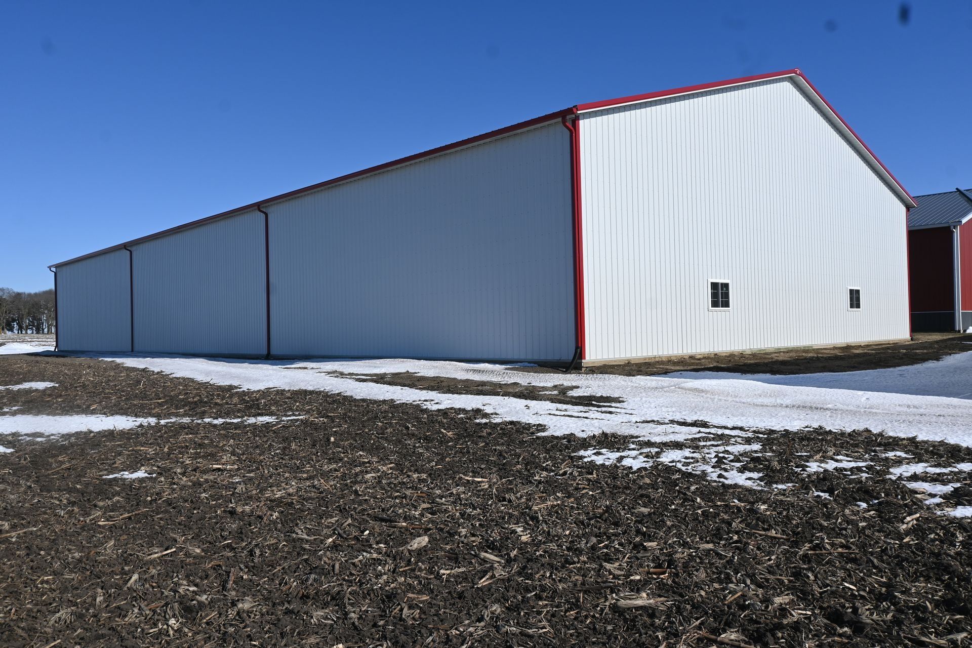White metal barn with red trim, windows, and roof on a field with snow and dark earth.