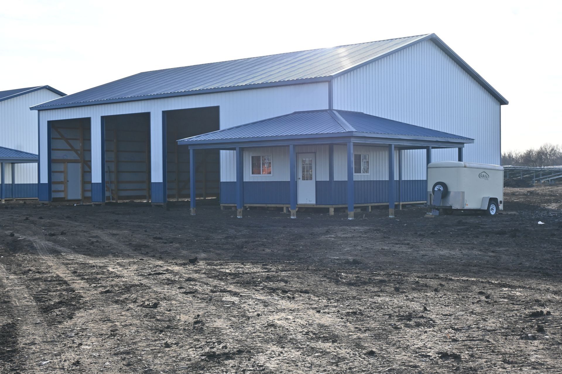 Blue and white metal building with attached porch on dirt lot, small trailer to the right.