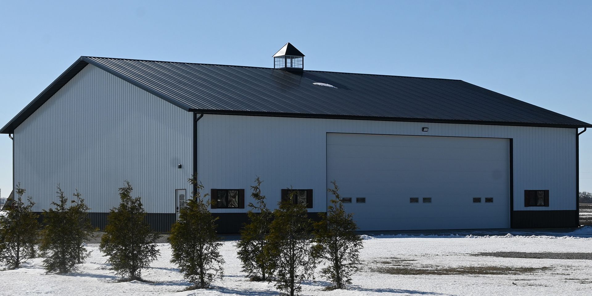 White and black metal barn with a black roof and a small decorative chimney on a snowy day.