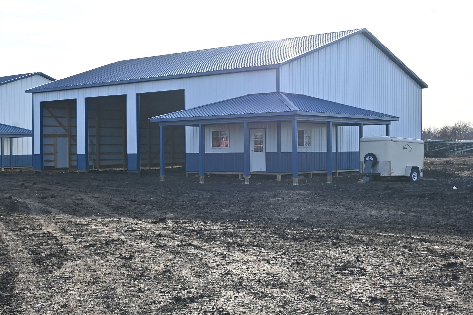 A blue and white metal building with an attached porch, on a dirt lot. A small trailer is parked nearby.