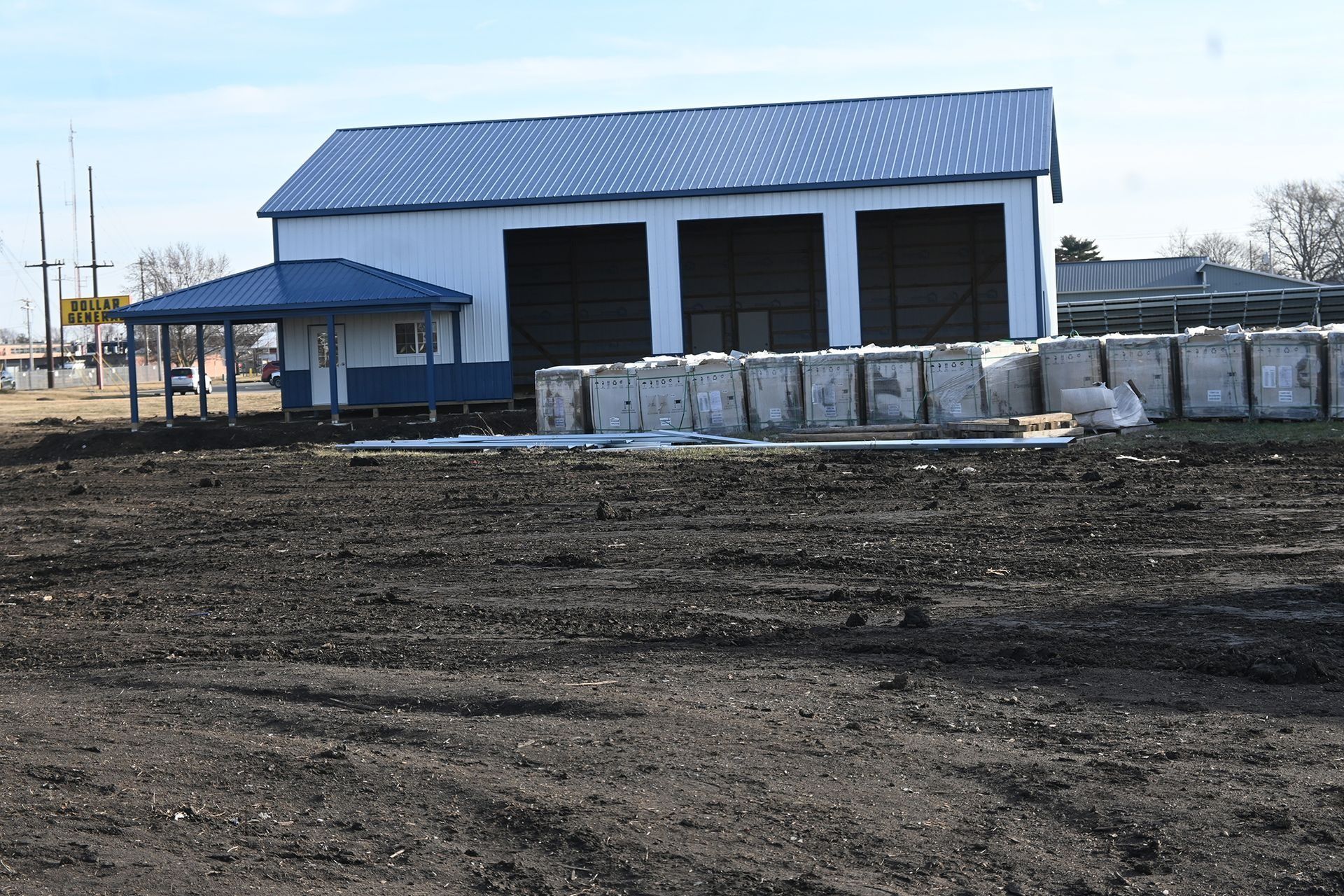 White building with blue roof and awning, dark bags stacked outside on a dirt lot.