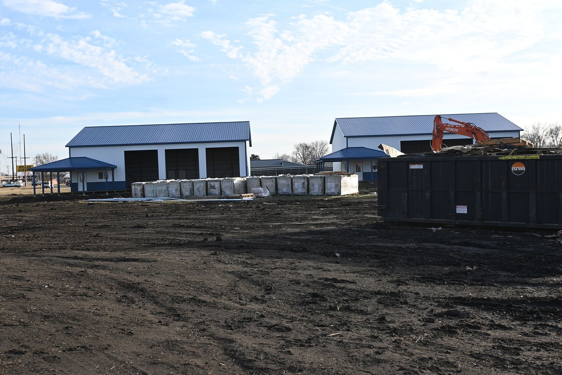 Construction site with two white buildings, blue roofs, and an excavator. Dark soil in the foreground.