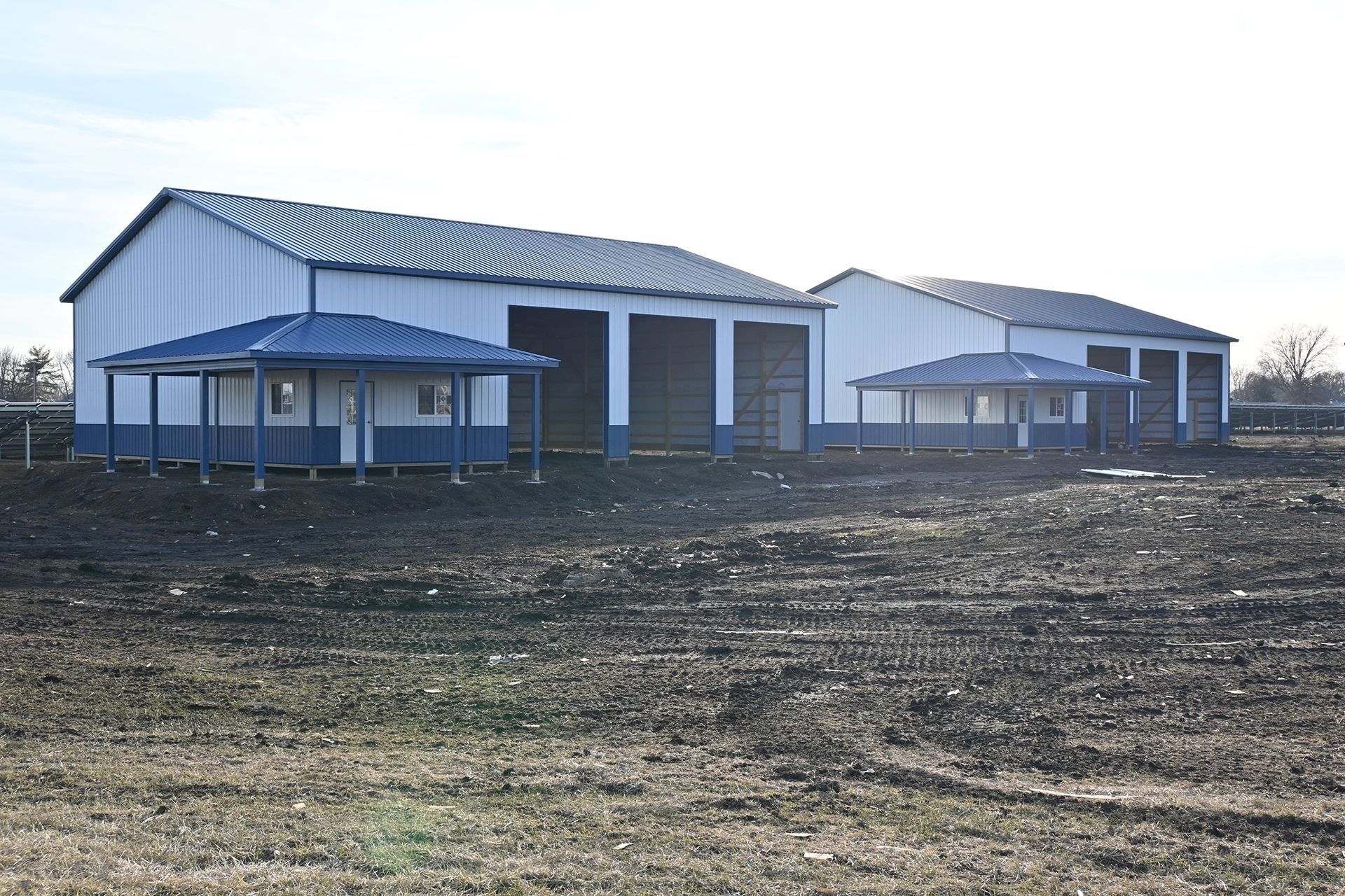 Two white metal buildings with blue trim, each with a blue-roofed porch, set in a brown field.